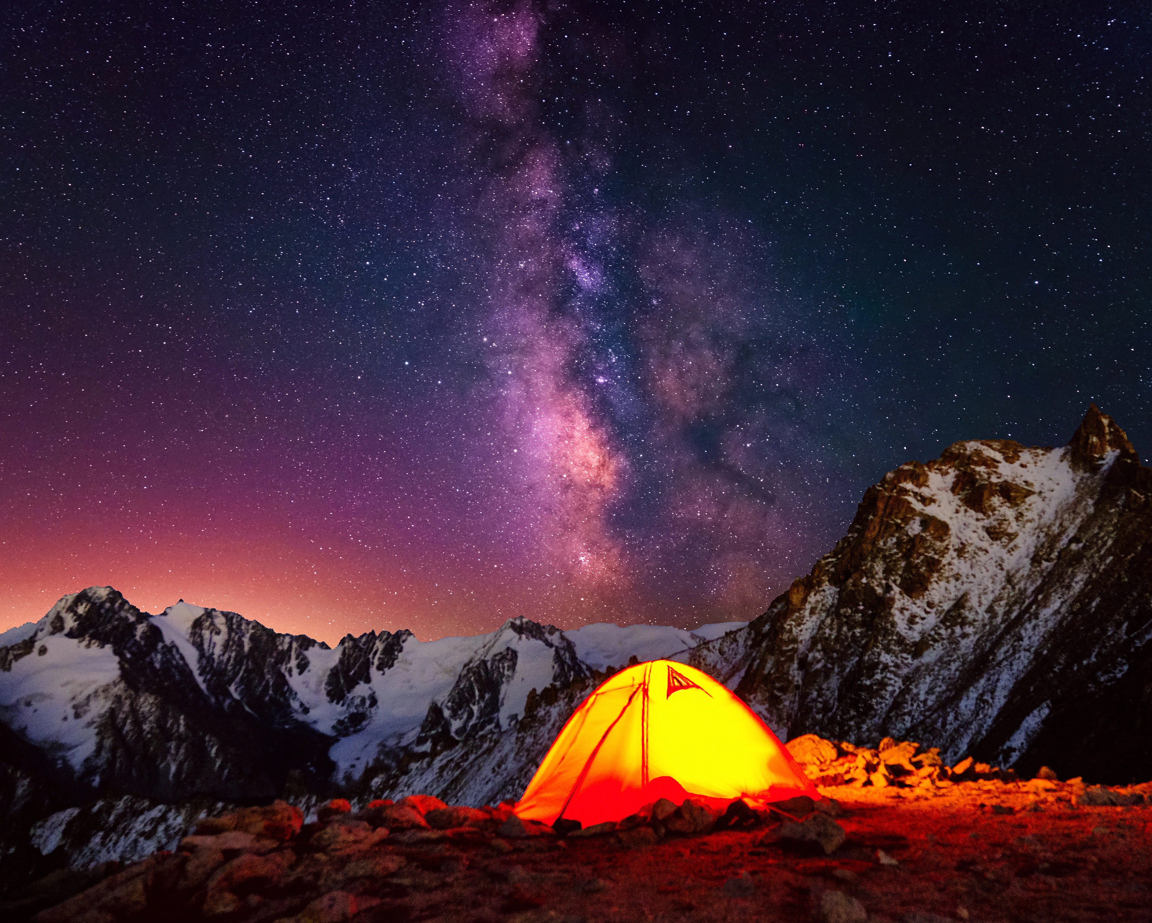 The Milky Way above snow-covered mountains and an illuminated tent.